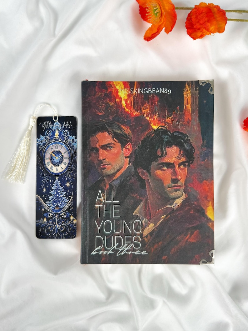 Op de afbeelding: Een boek getiteld "ALL THE YOUNG DUDES Book Three" met een omslag met twee mannen en een vurige achtergrond. Een bijpassende bladwijzer met een klokontwerp en kwast is ook aanwezig. De items staan op een witte stoffen achtergrond.