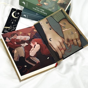 Op de afbeelding: Een stapel groene hardcover boeken getiteld "ALL THE YOUNG DUDES" met illustraties. Een open boek toont een gedetailleerde illustratie van twee handen met tatoeages en een rode draad. Een zwarte bladwijzer met een maan en sterren is ook zichtbaar.