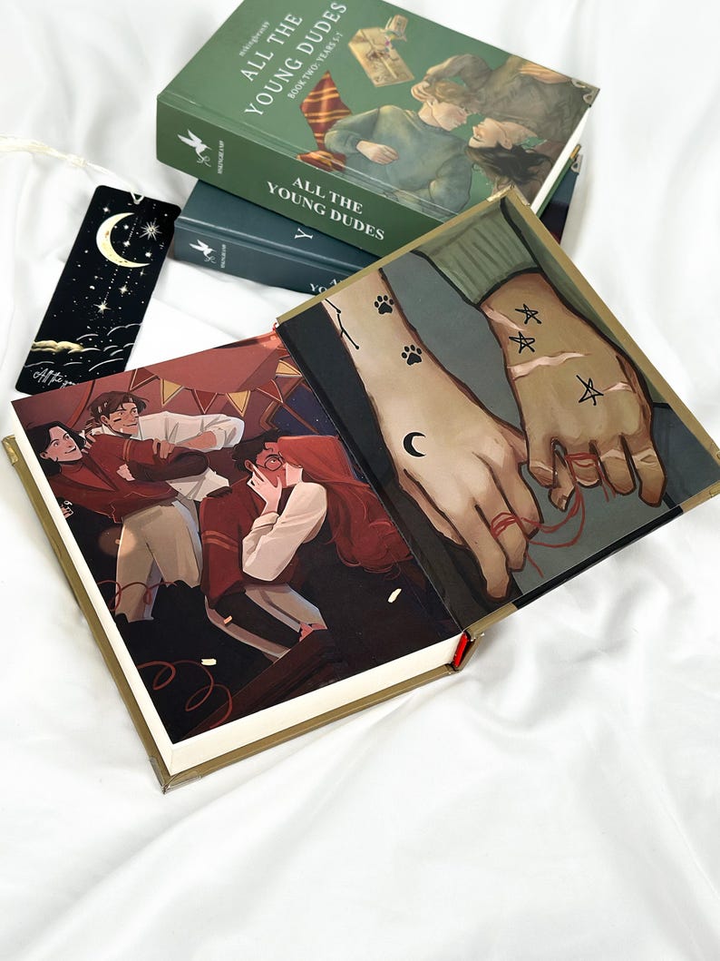 Op de afbeelding: Een stapel groene hardcover boeken getiteld "ALL THE YOUNG DUDES" met illustraties. Een open boek toont een gedetailleerde illustratie van twee handen met tatoeages en een rode draad. Een zwarte bladwijzer met een maan en sterren is ook zichtbaar.