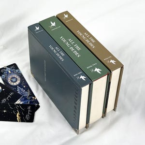 Op de afbeelding: Drie hardcover boeken getiteld "All The Young Dudes" in tinten blauw, groen en bruin, met witte tekst. De boeken zijn op elkaar gestapeld, met de ruggen naar boven. Bladwijzers met hemelse ontwerpen zijn zichtbaar.
