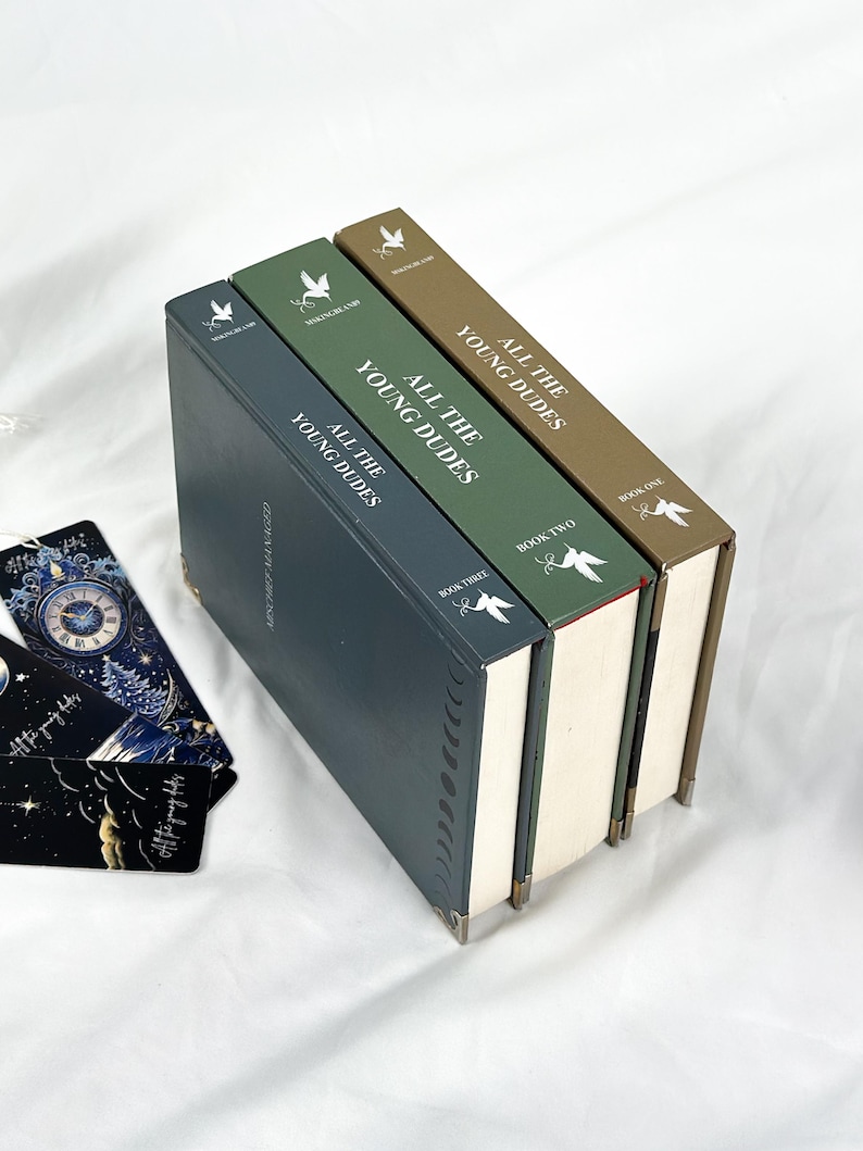 Op de afbeelding: Drie hardcover boeken getiteld "All The Young Dudes" in tinten blauw, groen en bruin, met witte tekst. De boeken zijn op elkaar gestapeld, met de ruggen naar boven. Bladwijzers met hemelse ontwerpen zijn zichtbaar.