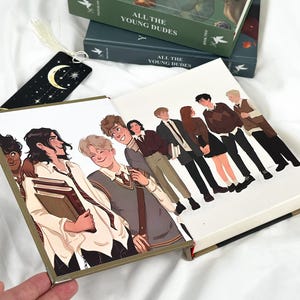 Op de afbeelding: Een stapel boeken getiteld "ALL THE YOUNG DUDES" met illustraties van jonge mensen. De boeken zijn groen en donkerblauw. Een zwarte bladwijzer met een maan en sterren is zichtbaar. De boekomslagen bevatten karakterillustraties.