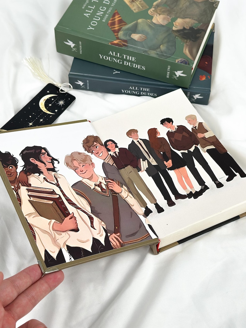 Op de afbeelding: Een stapel boeken getiteld "ALL THE YOUNG DUDES" met illustraties van jonge mensen. De boeken zijn groen en donkerblauw. Een zwarte bladwijzer met een maan en sterren is zichtbaar. De boekomslagen bevatten karakterillustraties.