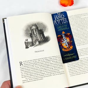 May include: A vertical rectangular bookmark featuring the title James Potter and the Curse of the Gatekeeper by G. Norman Lippert, displayed with a Gryffindor crest in red and gold, placed inside an open book with a printed prologue page.