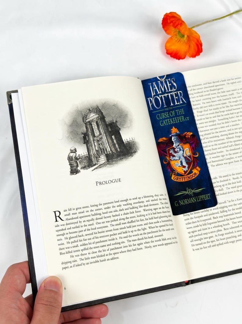 May include: A vertical rectangular bookmark featuring the title James Potter and the Curse of the Gatekeeper by G. Norman Lippert, displayed with a Gryffindor crest in red and gold, placed inside an open book with a printed prologue page.