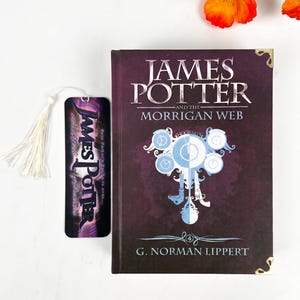 May include: A hardcover book titled James Potter and the Morrigan Web by G. Norman Lippert with a dark purple cover, gold corner accents, and a central clock graphic, accompanied by a matching vertical bookmark with a white tassel.