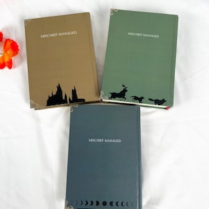 Op de afbeelding: Drie hardcover boeken met de tekst "MISCHIEF MANAGED". De boeken zijn in tinten bruin, groen en blauwgrijs. Elk boek heeft een silhouetontwerp: een kasteel, een hert met honden en een reeks halve manen.