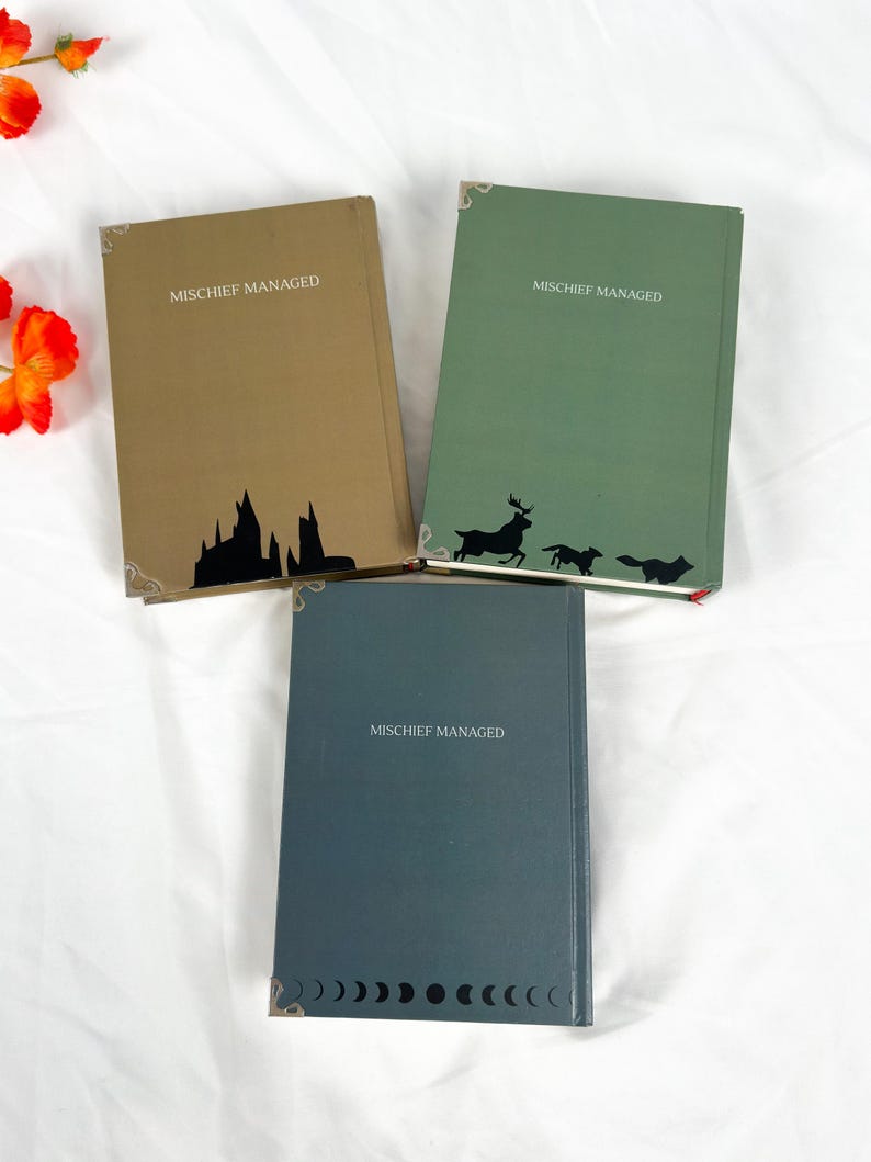 Op de afbeelding: Drie hardcover boeken met de tekst "MISCHIEF MANAGED". De boeken zijn in tinten bruin, groen en blauwgrijs. Elk boek heeft een silhouetontwerp: een kasteel, een hert met honden en een reeks halve manen.