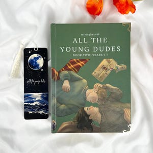 Op de afbeelding: Een groen boek met de titel "ALL THE YOUNG DUDES BOOK TWO: YEARS 5-7" met een illustratie van twee personen. Een zwarte bladwijzer met een maan- en oceaanontwerp en de tekst "All the young dudes" staat naast het boek. Oranje bloemen staan op de achtergrond.