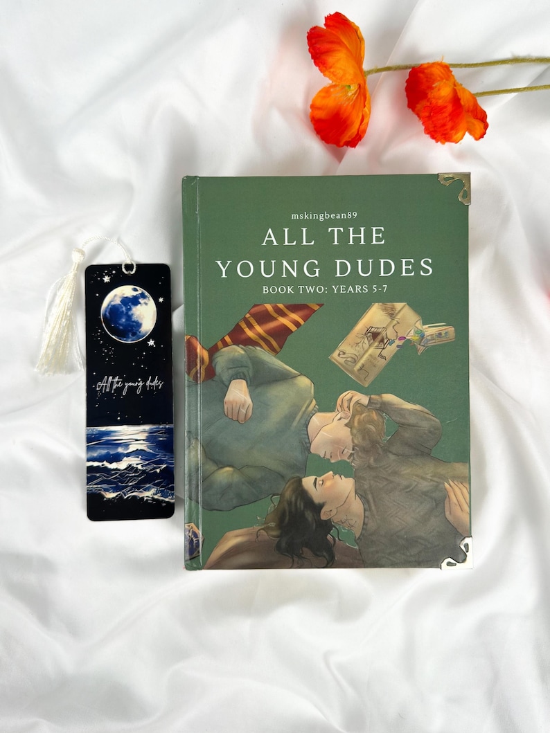 Op de afbeelding: Een groen boek met de titel "ALL THE YOUNG DUDES BOOK TWO: YEARS 5-7" met een illustratie van twee personen. Een zwarte bladwijzer met een maan- en oceaanontwerp en de tekst "All the young dudes" staat naast het boek. Oranje bloemen staan op de achtergrond.