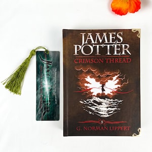 May include: A hardcover book titled James Potter and the Crimson Thread by G. Norman Lippert with a dark brown cover featuring a silhouette against a bright light and waves, alongside a matching rectangular bookmark with a green tassel.