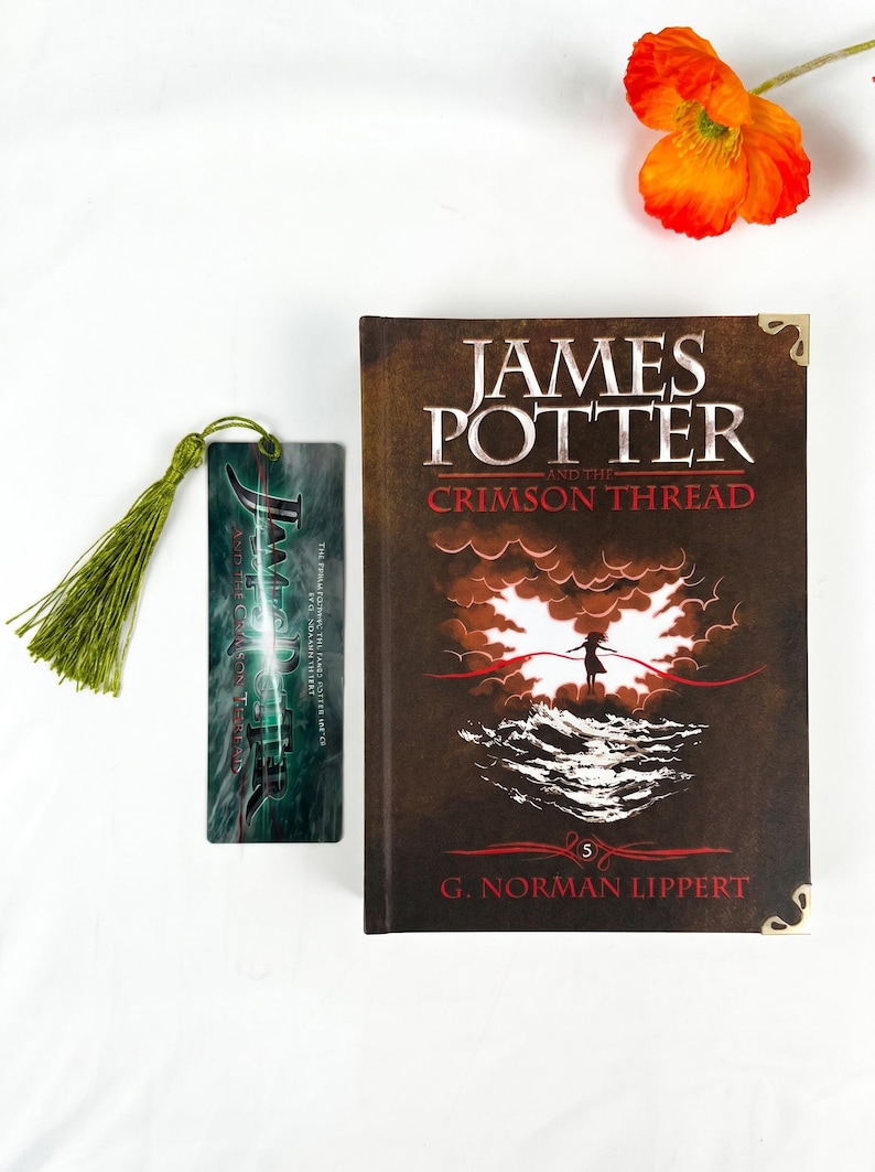 May include: A hardcover book titled James Potter and the Crimson Thread by G. Norman Lippert with a dark brown cover featuring a silhouette against a bright light and waves, alongside a matching rectangular bookmark with a green tassel.