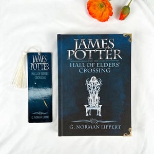 May include: A hardcover book titled James Potter and the Hall of Elders' Crossing by G. Norman Lippert with a dark blue cover featuring a white throne illustration, alongside a matching rectangular bookmark with a white tassel.