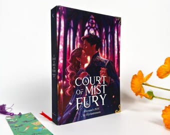 Könnte beinhalten: Ein Hardcover-Buch mit dem Titel "Court of Mist and Fury" mit einem Lesezeichen. Das Buchcover zeigt ein sich umarmendes Paar in einer dunklen, verzierten Umgebung. Das Lesezeichen ist grün mit Illustrationen und Text.