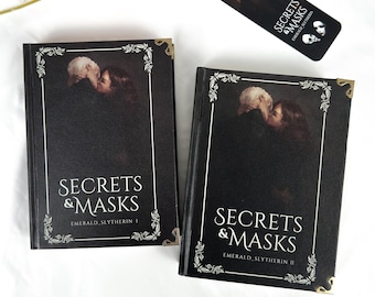 Könnte beinhalten: Zwei schwarze Hardcover-Bücher und ein Lesezeichen mit dem Titel "Secrets & Masks". Die Bücher haben goldene Eckakzente und einen dekorativen Rand. Das Lesezeichen hat eine goldene Quaste und ein passendes Design.