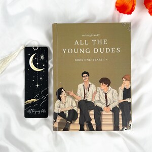 Op de afbeelding: Een boek getiteld "ALL THE YOUNG DUDES" met een illustratie van vier jonge mannen op de cover. Een zwarte bladwijzer met een maan- en sterrenontwerp en de tekst "All the young dudes" staat naast het boek. Oranje bloemen staan op de achtergrond.