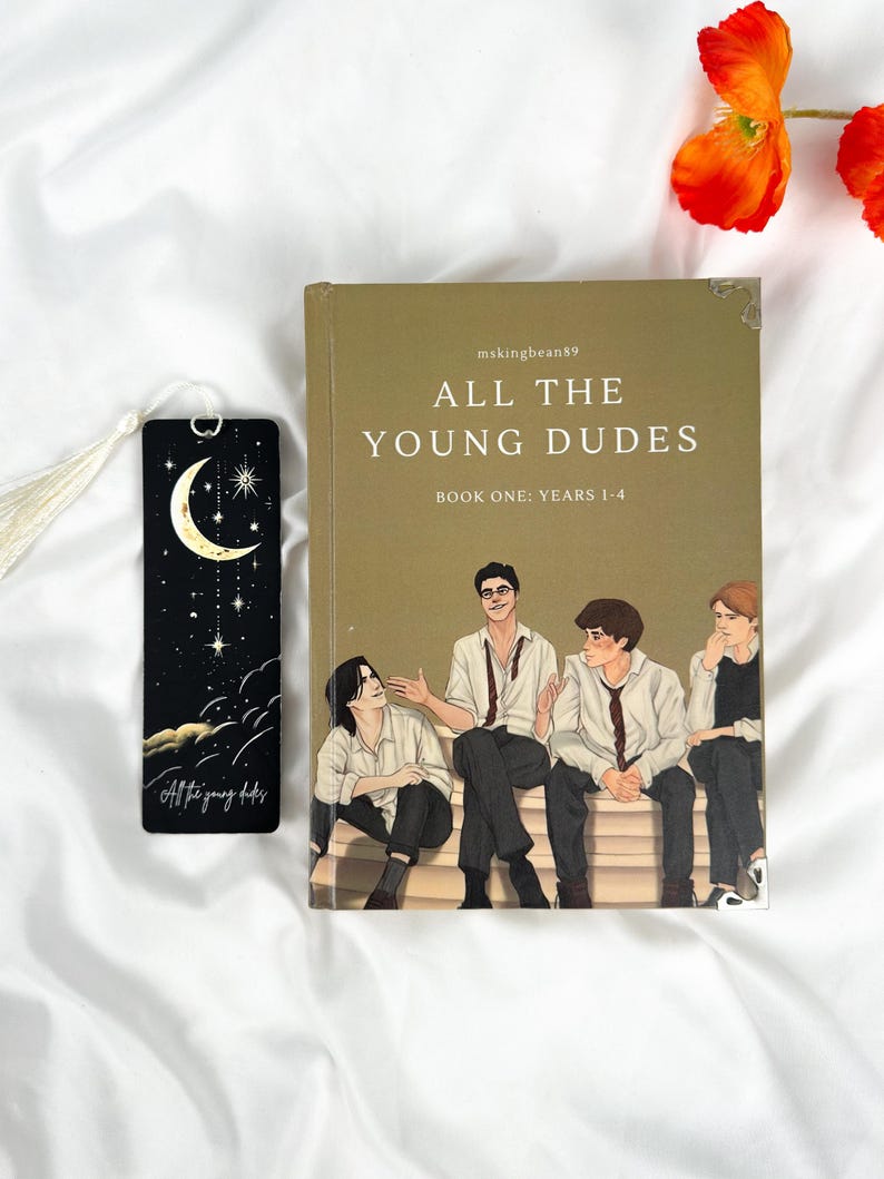 Op de afbeelding: Een boek getiteld "ALL THE YOUNG DUDES" met een illustratie van vier jonge mannen op de cover. Een zwarte bladwijzer met een maan- en sterrenontwerp en de tekst "All the young dudes" staat naast het boek. Oranje bloemen staan op de achtergrond.