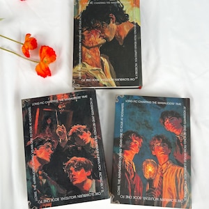 Op de afbeelding: Drie boeken met illustraties van personages. Het bovenste boek toont twee kussende mensen. De andere twee boeken hebben illustraties van jonge mannen. De boeken liggen op een wit oppervlak met oranje bloemen.
