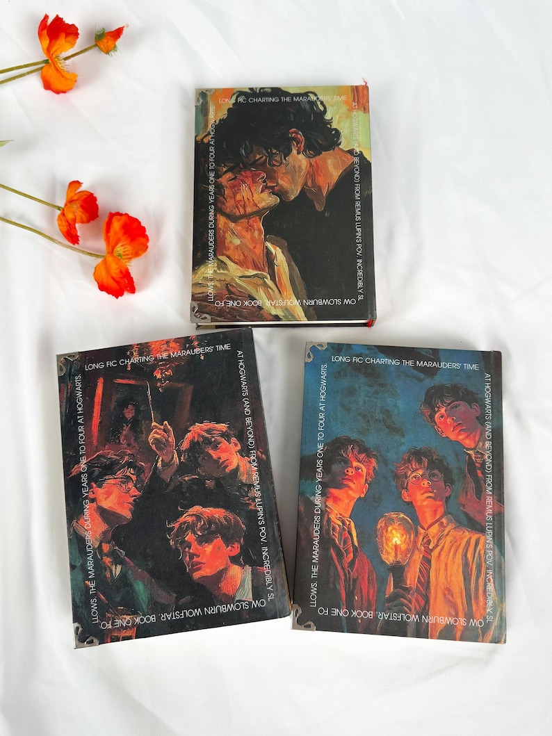 Op de afbeelding: Drie boeken met illustraties van personages. Het bovenste boek toont twee kussende mensen. De andere twee boeken hebben illustraties van jonge mannen. De boeken liggen op een wit oppervlak met oranje bloemen.