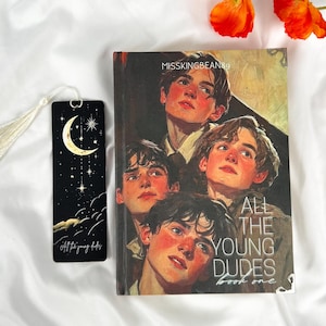 Op de afbeelding: Een boek- en bladwijzerset. De boekcover toont een portret van vijf jonge mannen. De bladwijzer is zwart met een maan- en sterrenontwerp. De tekst "All the young dudes book one" is zichtbaar op het boek.