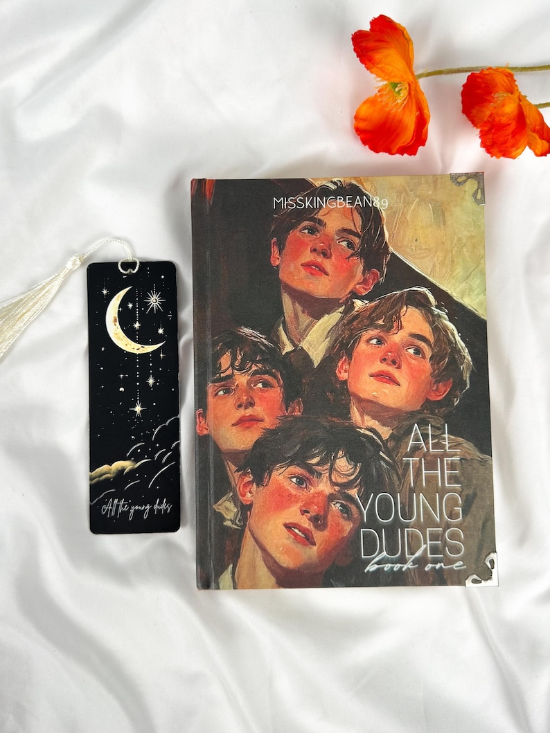Op de afbeelding: Een boek- en bladwijzerset. De boekcover toont een portret van vijf jonge mannen. De bladwijzer is zwart met een maan- en sterrenontwerp. De tekst "All the young dudes book one" is zichtbaar op het boek.