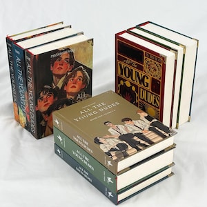 Op de afbeelding: Verschillende hardcover boeken met de titel "All The Young Dudes". De boeken hebben verschillende omslagontwerpen, waaronder portretten van jonge mensen en gestileerde tekst. De ruggen tonen de titel en de volumenummers.