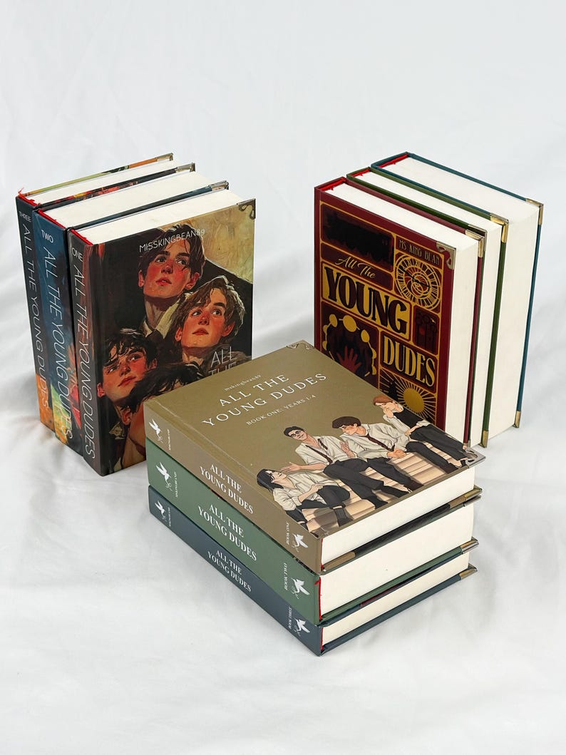 Op de afbeelding: Verschillende hardcover boeken met de titel "All The Young Dudes". De boeken hebben verschillende omslagontwerpen, waaronder portretten van jonge mensen en gestileerde tekst. De ruggen tonen de titel en de volumenummers.