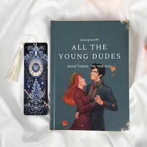 Op de afbeelding: Een boek getiteld "ALL THE YOUNG DUDES" met een omslagillustratie van een dansend stel. Een bijpassende bladwijzer met een klokontwerp en kwast ligt naast het boek. Het boek en de bladwijzer liggen op een witte stoffen achtergrond.