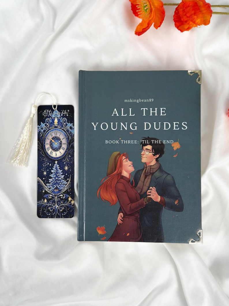 Op de afbeelding: Een boek getiteld "ALL THE YOUNG DUDES" met een omslagillustratie van een dansend stel. Een bijpassende bladwijzer met een klokontwerp en kwast ligt naast het boek. Het boek en de bladwijzer liggen op een witte stoffen achtergrond.