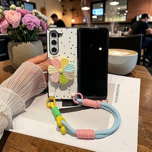 Peut inclure: Coque de téléphone blanche avec un motif floral et un accent de fleur multicolore. L'étui a une sangle avec un cordon bleu et rose et un nœud jaune et vert. Le téléphone est ouvert, révélant un écran noir.