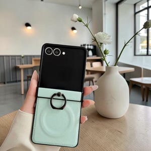 Può includere: Una custodia per telefono nera e verde menta con un anello di supporto. La custodia è aperta, rivelando la fotocamera e lo schermo del telefono. La sezione verde menta ha un design circolare. La custodia è tenuta in mano.