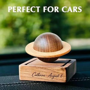 May include: A wooden planet-shaped ornament with a dark brown sphere and a light tan ring, mounted on a square wooden base. The base features the engraved text 'Cathrine · August 8' and the image includes the white text 'PERFECT FOR CARS'.