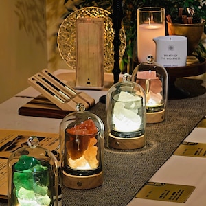 Puede incluir: Tres lámparas de cúpula de cristal con bases de madera, cada una con piedras minerales en bruto iluminadas en verde, naranja y blanco. Las lámparas se sitúan sobre una mesa junto a una vela blanca con la etiqueta Breath of the Wilderness.