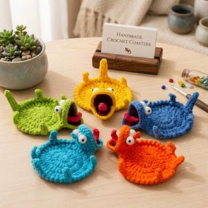 Peut inclure: Cinq dessous de verre ronds au crochet en forme de monstres colorés avec des bouches ouvertes, des langues et des yeux mobiles. Les dessous de verre sont vert citron, jaune, bleu, bleu sarcelle et orange. Un porte-carte affiche Handmade Crochet Coasters.