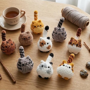 May include: A collection of handmade crochet cat toys in various colors, including orange, brown, gray, and white, with detailed features. The scene includes a cup of tea, a succulent plant, and craft supplies, all arranged on a wooden surface.