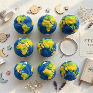 May include: Nine blue, yellow, and green crocheted Earth globes are arranged on a light surface. Decorative wooden planets, stars, and moons surround the globes. A small notebook, pen, and a card with text are also present. The theme is educational and decorative.