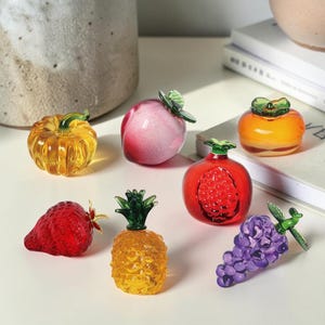 Könnte beinhalten: Eine Sammlung farbenfroher Glasfruchtskulpturen, darunter ein Kürbis, ein Pfirsich, eine Erdbeere, eine Ananas, ein Granatapfel, eine Kaki und Trauben. Die Glasfruchtstücke sind in verschiedenen Orange-, Rot-, Gelb-, Rosa- und Lilatönen gehalten.