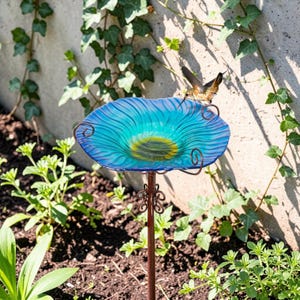 Puede incluir: Un bebedero para pájaros de vidrio azul con un centro amarillo, sostenido por un soporte de metal decorativo. El bebedero tiene una textura ondulada y está en un jardín con plantas verdes y un colibrí posado en el borde.