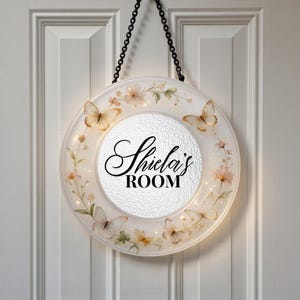 May include: A round, illuminated sign with the text "Shiela's ROOM" in elegant script. The sign features butterfly and floral designs in soft colors, with a chain for hanging. The background is a white door.