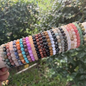 Könnte beinhalten: Eine Sammlung von Perlenarmbändern in verschiedenen Farben, darunter Rosa, Blau, Braun und Grün. Die Armbänder werden auf einem weißen zylindrischen Halter mit goldenem Rand vor grünem Laubwerk präsentiert.