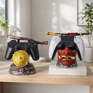 May include: Two gaming controller stands. One features a black controller with an axe and a gold disc. The other has a white and black controller with a red and gold mask and a katana. Both are on a wooden surface.
