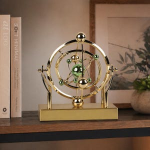 May include: A gold-colored, decorative kinetic sculpture with a spherical green center and orbiting spheres. The sculpture is mounted on a rectangular base and features multiple circular and curved elements. It is displayed on a wooden surface.