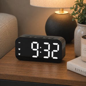 May include: A black digital alarm clock displaying 9:32 AM with bright white numerals. The clock has a rectangular shape and sits on a wooden surface. The clock is next to a lamp and decorative items.