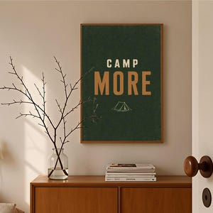 May include: A rectangular wall art print in a wooden frame featuring a dark green background with the text CAMP MORE in bold tan letters and a small white line drawing of a tent centered below the text, displayed in a clean interior setting.