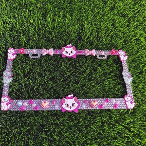 May include: A pink license plate frame decorated with rhinestones, bows, and cat-shaped embellishments. The frame features a repeating pattern of pink and clear rhinestones, with cat and bow accents. The frame is set against a green background.