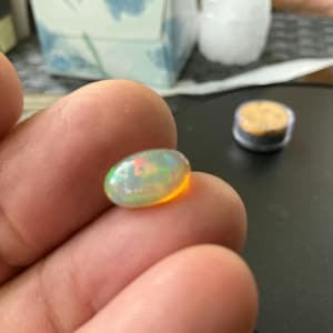 May include: An oval-shaped opal gemstone with a vibrant play of color, including green, yellow, and hints of red. The gemstone is held in a hand, showcasing its iridescent qualities. A small, round container is in the background.