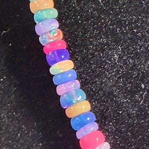 May include: A colorful beaded necklace featuring a variety of opal beads in shades of pink, blue, yellow, and purple. The beads are oval-shaped and strung together, creating a vibrant and eye-catching accessory.