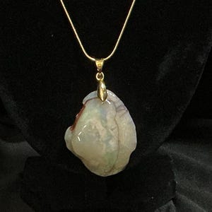 Könnte beinhalten: Eine goldfarbene Halskette mit Anhänger. Der Anhänger ist ein großer, unregelmäßig geformter Stein mit einer Mischung aus Farben, darunter Weiß, Lila und Andeutungen von Grün und Rot. Der Stein ist der Mittelpunkt der Halskette.