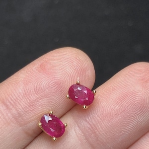 May include: A pair of stud earrings featuring oval-cut, deep pinkish-red rubies set in yellow gold prongs. Each gemstone displays a faceted surface that reflects light, secured by four small gold prongs at the cardinal points of the oval shape.