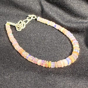 May include: A delicate bracelet featuring small, iridescent beads in shades of pink, orange, and blue. The beads are strung together with a silver-colored chain and clasp. The bracelet is displayed against a dark background.
